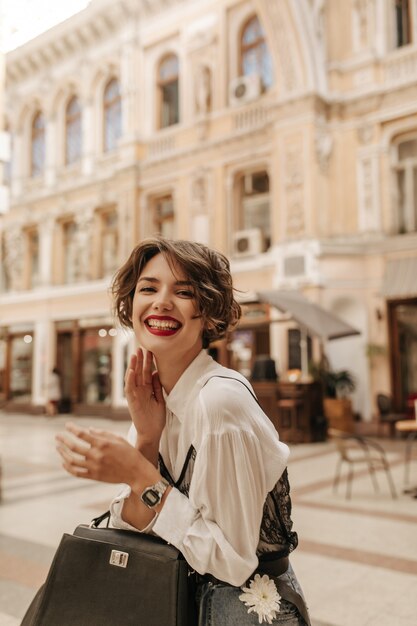 Femme positive avec des lèvres rouge vif et des cheveux ondulés en riant dans la ville. Femme cool en chemise blanche avec sac à main posant dans la rue.