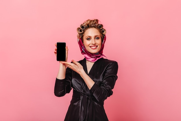 Femme positive en robe noire et écharpe démontre smartphone sur mur rose