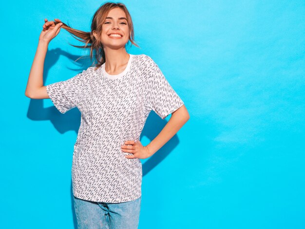 Femme positive souriant. Modèle drôle posant près du mur bleu en studio, elle touche ses cheveux