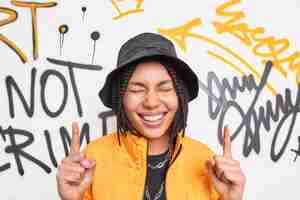 Photo gratuite femme positive sourit largement au-dessus avec deux index a une expression heureuse vêtue de vêtements à la mode pose contre le mur de graffitis