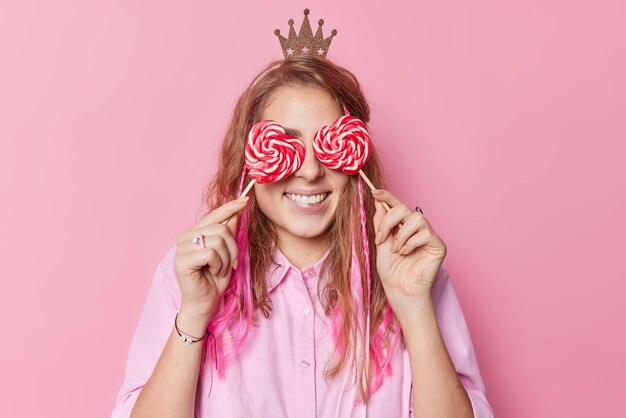 Une femme positive sourit largement couvre les yeux avec des bonbons en forme de coeur porte une petite couronne a les cheveux longs étant de bonne humeur pose contre un fond rose aime un dessert sucré Goûtez deux sucettes