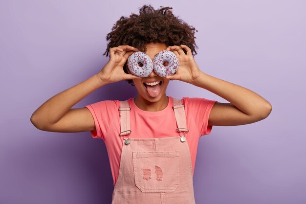 La femme positive tient deux beignets violets sur les yeux, sort la langue, porte un t-shirt rose et une salopette, étant gourmande, s'amuse, pose à l'intérieur sur un mur violet. Mange un dessert savoureux avec moi