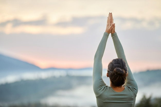 Une femme pratiquant le yoga fait la pose du guerrier devant des montagnes brumeuses le matin.