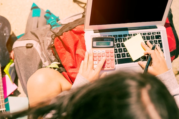 Femme prenant des notes à la main avec la calculatrice sur un ordinateur portable en journée ensoleillée
