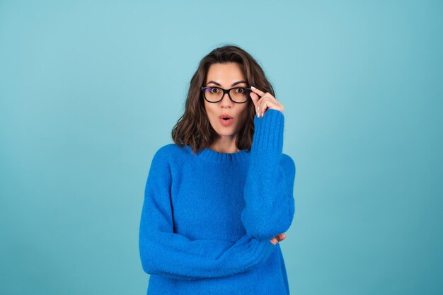 Femme en pull bleu et maquillage naturel, cheveux courts bouclés, lunettes sur les yeux, choquée, surprise avec la bouche ouverte