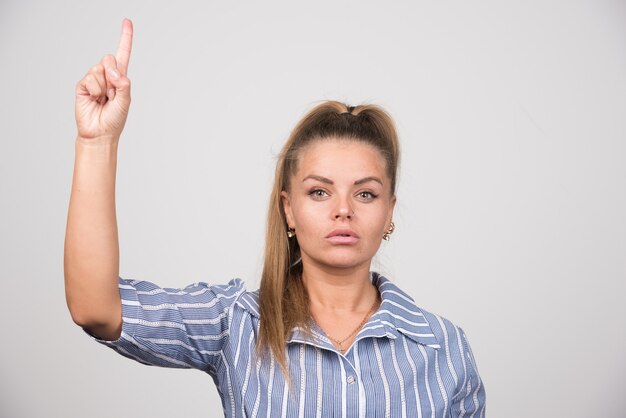 Femme en pull bleu pointant vers le haut sur un mur gris.