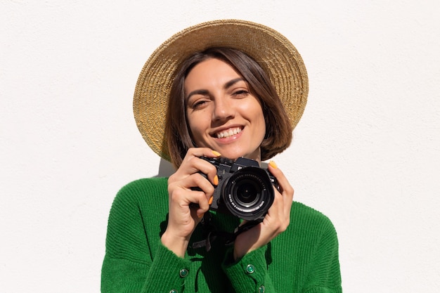 Femme en pull décontracté vert et chapeau en plein air sur mur blanc heureux touriste positif avec appareil photo professionnel