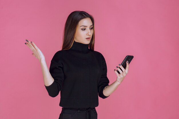 Femme en pull noir tenant un smartphone et envoyer des SMS ou vérifier les médias sociaux.