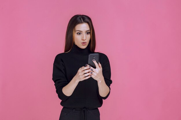 Femme en pull noir tenant un smartphone et envoyer des SMS ou vérifier les médias sociaux.