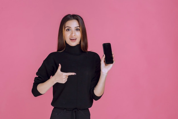 Femme en pull noir tenant un smartphone et pointant dessus.