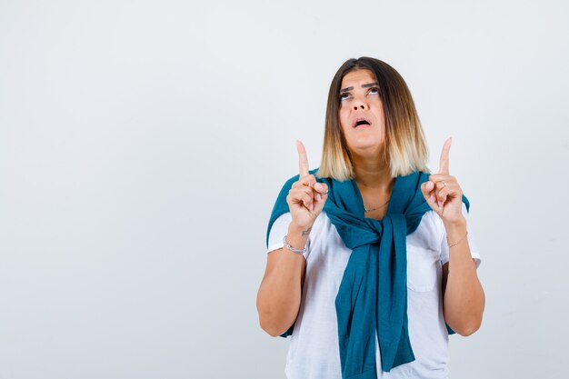 Femme avec pull noué pointant vers le haut, regardant vers le haut en t-shirt blanc et ayant l'air plein d'espoir, vue de face.