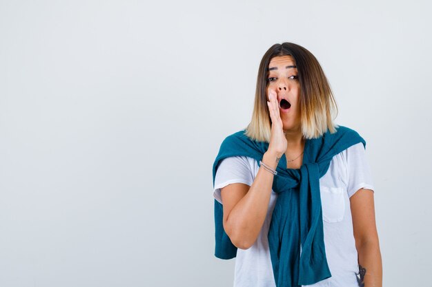 Femme avec pull noué en t-shirt blanc gardant la main près de la bouche tout en racontant le secret et l'air excité, vue de face.