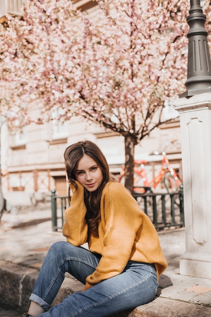 Femme en pull orange est assise sur la route de la rue. Charmante jeune fille en jeans posant à l'extérieur près de bloomimg sakura. Dame regardant dans la caméra