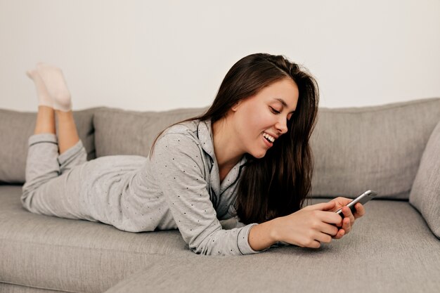 Femme en pyjama allongé sur le canapé avec smartphone et défilement. Fille européenne au repos à la maison.