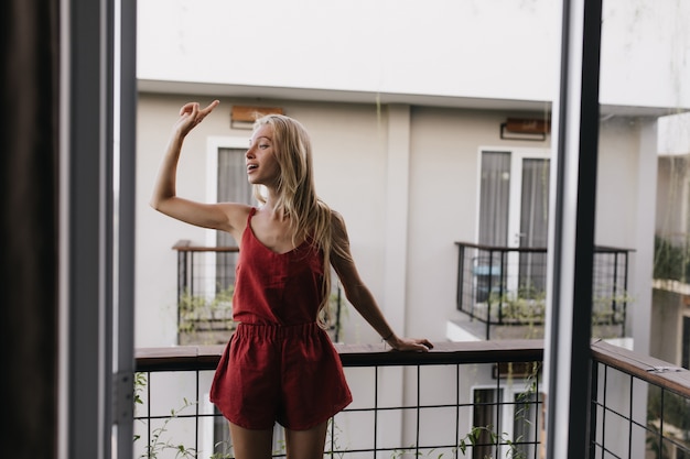 femme en pyjama debout au balcon et regardant autour. Modèle féminin excité avec de longs cheveux blonds appréciant le matin.