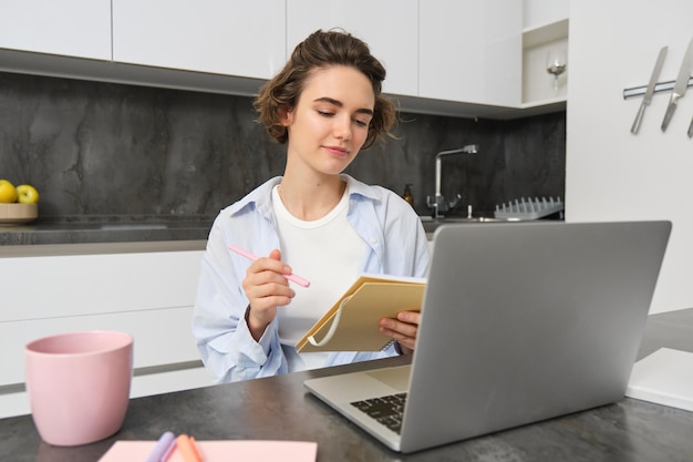 Une femme qui travaille prend des notes et écrit des informations sur un ordinateur portable assis avec un ordinateur portable à la maison