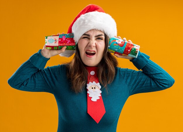 Femme de race blanche adulte ennuyé avec Bonnet de Noel et cravate de Père Noël tenant des gobelets en papier près des oreilles isolé sur fond orange avec espace de copie