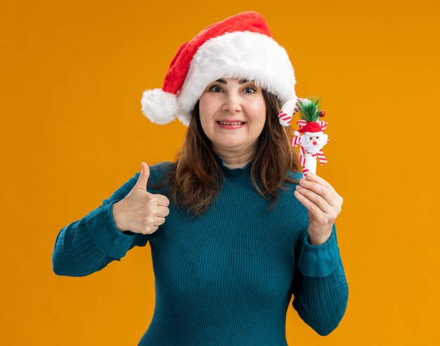 Femme de race blanche adulte impressionnée avec bonnet de Noel détient la canne en bonbon et les pouces vers le haut isolé sur fond orange avec copie espace