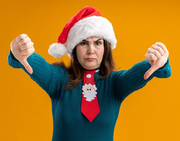 Femme de race blanche adulte non satisfaite avec Bonnet de Noel et cravate de père Noël pouces vers le bas isolé sur fond orange avec espace de copie