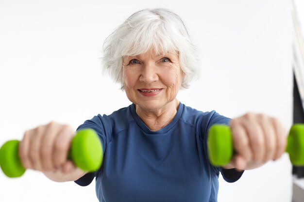 Femme de race blanche âgée heureuse énergique active aux cheveux gris bénéficiant d'exercices physiques à l'intérieur, s'entraînant à la maison à l'aide d'haltères, souriant largement. Mise au point sélective sur le visage de la femme