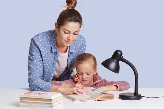 Femme de race blanche aidant son doughter à faire ses devoirs, mère et enfant entourés de livres, petite fille assise concentrée au bureau blanc, essayant de faire des sommes. Concept de l'éducation.