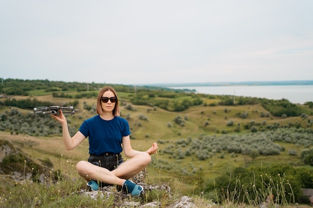 Une femme de race blanche avec un drone à la main, assis sur une colline rocheuse verte avec ciel