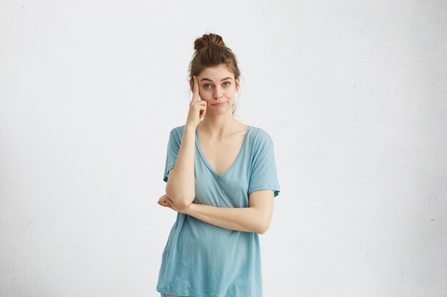 Femme de race blanche réfléchie portant un t-shirt bleu décontracté tenant le doigt sur sa tempe, ayant confus un regard pensif, réfléchissant, pesant tous les avantages et inconvénients de la proposition