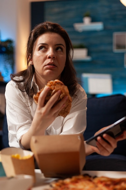 femme de race blanche tenant un hamburger tastu dans les mains parcourant les médias sociaux à l'aide d'un smartphone