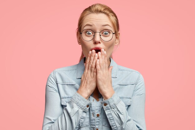 Femme à la recherche agréable et émotionnelle d'apparence européenne, glousse à quelque chose, couvre la bouche avec les deux paumes, a une expression étonnée, porte des lunettes et une veste en jean, isolée sur un mur rose