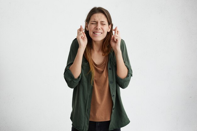 Femme réconfortante d'espoir gardant ses doigts croisés et les yeux fermés fronçant les sourcils son visage ayant un grand espoir et souhaitant prier pour bonne chance