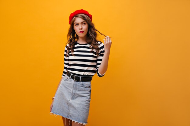 Femme réfléchie en béret rouge touchant ses cheveux. Jolie fille aux cheveux ondulés en jupe en jean avec ceinture noire posant sur fond orange.