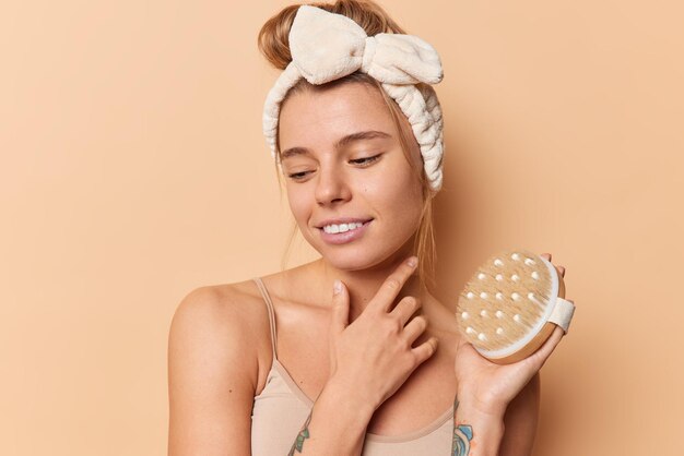 Une femme réfléchie concentrée sur le visage tient doucement une brosse sèche pour exfolier la peau porte un bandeau et un t-shirt isolés sur fond beige. Traitement de la cellulite et concept de brossage à sec