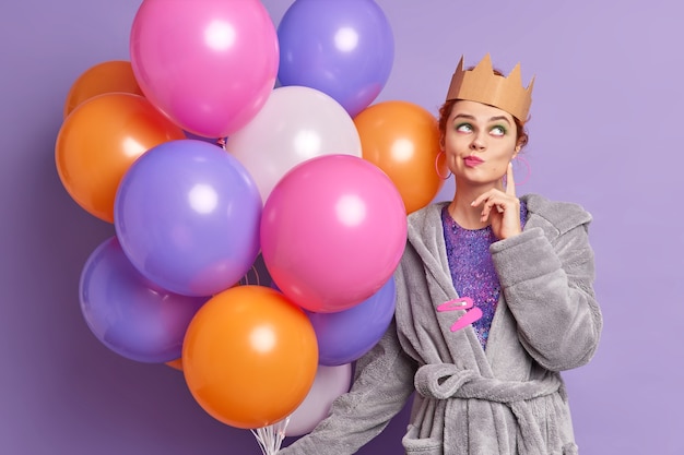 Une femme réfléchie a l'image de la reine porte une couronne sur la tête se dresse pensif concentré au-dessus des lèvres des sacs à main pense à la célébration des vacances à venir détient des ballons gonflés multicolores