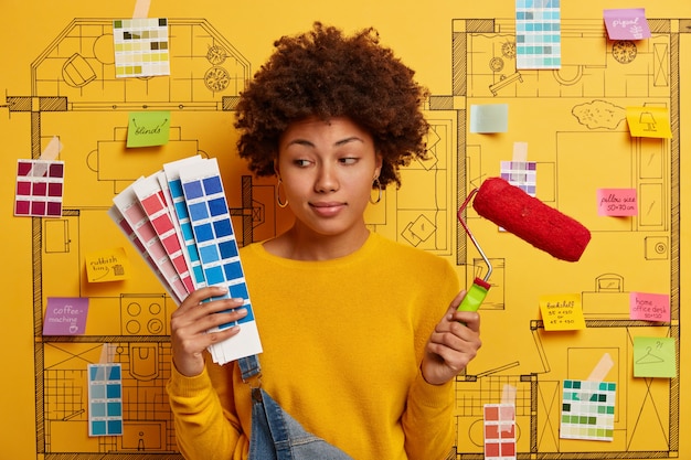 Une femme réfléchie et indécise regarde des échantillons de couleurs, tient un rouleau à peinture, pense à la rénovation des murs d'une nouvelle maison, pose contre un croquis avec des notes écrites autocollantes. Réparation, construction, concept de maison