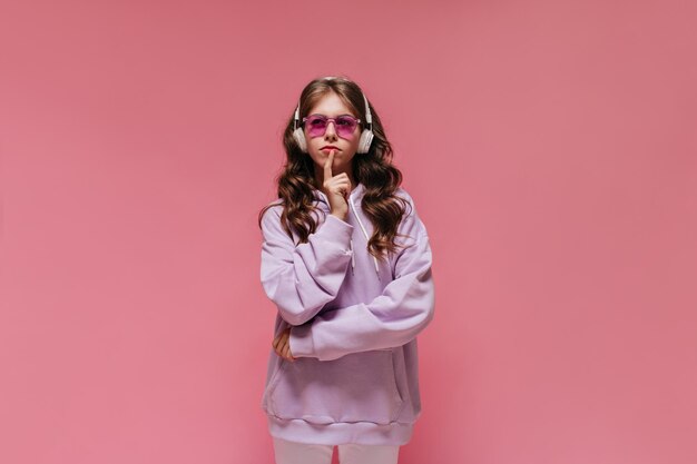 Femme réfléchie en sweat à capuche violet et pantalon blanc pose sur une fille brune isolée écoutant de la musique dans des écouteurs sur fond rose