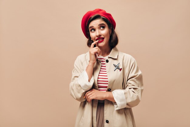 Femme réfléchie en tenue beige pose sur fond isolé. Fille élégante avec rouge à lèvres brillant en béret rouge et en manteau lève les yeux.