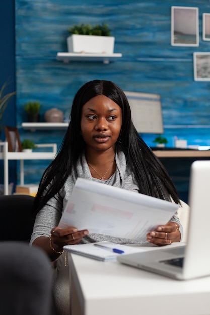 Femme regardant un écran d'ordinateur portable et tenant des papiers avec des graphiques de données, faisant du travail à distance. Jeune adulte travaillant à domicile sur un projet d'entreprise tout en utilisant un ordinateur et en concevant des fichiers.