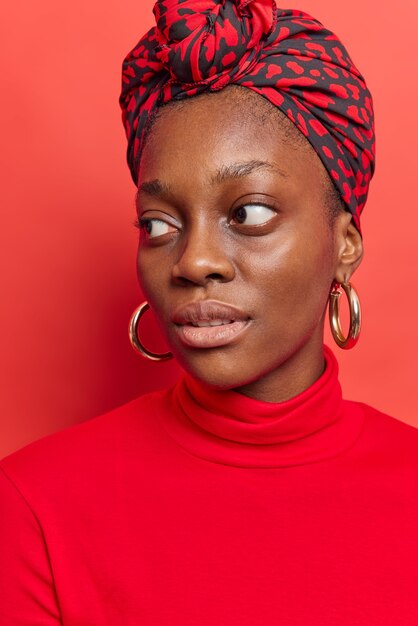 la femme regarde au loin avec une expression réfléchie porte un foulard à col roulé noué sur la tête de grosses boucles d'oreilles pose sur un rouge vif