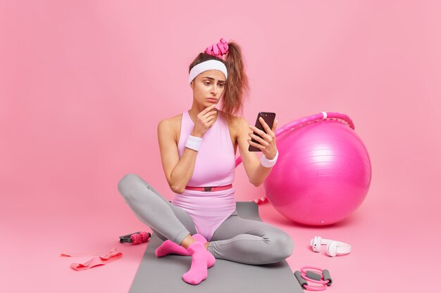 la femme regarde l'écran du smartphone a une silhouette sportive vérifie les messages ou le fil d'actualité dans les réseaux sociaux est assise sur un tapis confortable