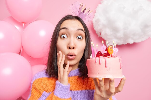 la femme regarde se demande à l'appareil photo tient un délicieux gâteau d'anniversaire savoureux porte une couronne et des poses de cavalier rayé