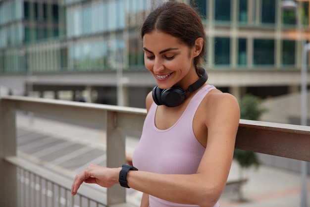 une femme regarde une smartwatch heureuse de brûler beaucoup de calories pendant l'entraînement porte des écouteurs courts autour du cou se tient régulièrement en plein air