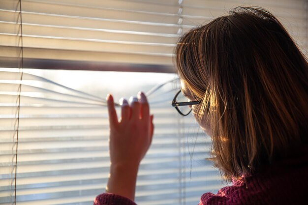 Une femme regarde à travers les stores à la lumière du soleil tôt le matin