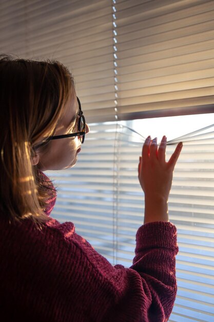 Une femme regarde à travers les stores à la lumière du soleil tôt le matin