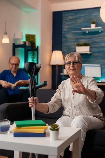 Femme à la retraite avec des béquilles ondulant lors d'une vidéoconférence à la maison. Femme plus âgée utilisant la communication à distance en ligne tandis que son mari handicapé est assis en fauteuil roulant sur fond de salon