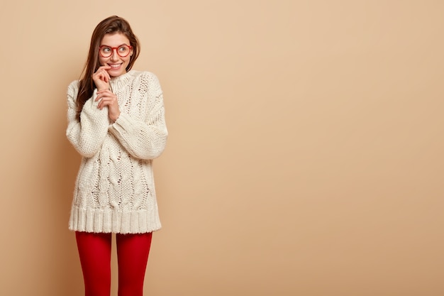 Une femme rêveuse heureuse a les cheveux raides, a l'air pleinement heureuse avec une expression réfléchie, imagine un moment positif dans la vie, porte un pull blanc, des leggings rouges, isolé sur un mur beige avec un espace libre