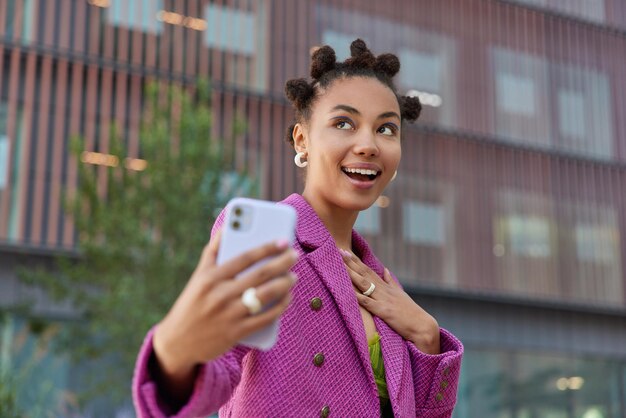 Une femme rêveuse heureuse avec une coiffure amusante fait une photo d'elle-même porte une veste rose à la mode et pose positivement à l'extérieur sur un arrière-plan flou. Technologie et style de vie urbain