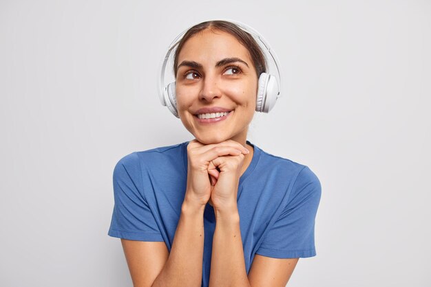 Une femme rêveuse optimiste garde les mains sous le menton regarde au loin écoute de la musique dans des écouteurs stéréo et a des pensées agréables porte un t-shirt bleu décontracté isolé sur un mur blanc