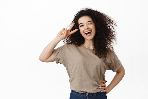 femme, riant et souriant, montrant le signe v de la paix près de l'œil, pose kawaii, debout sur blanc