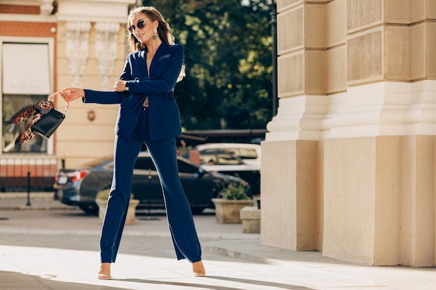 Femme riche de luxe vêtue d'un élégant costume bleu élégant marchant dans la ville aux beaux jours d'automne tenant le sac à main