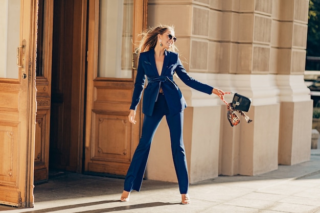 Femme riche de luxe vêtue d'un élégant costume bleu élégant marchant dans la ville aux beaux jours d'été tenant sac à main